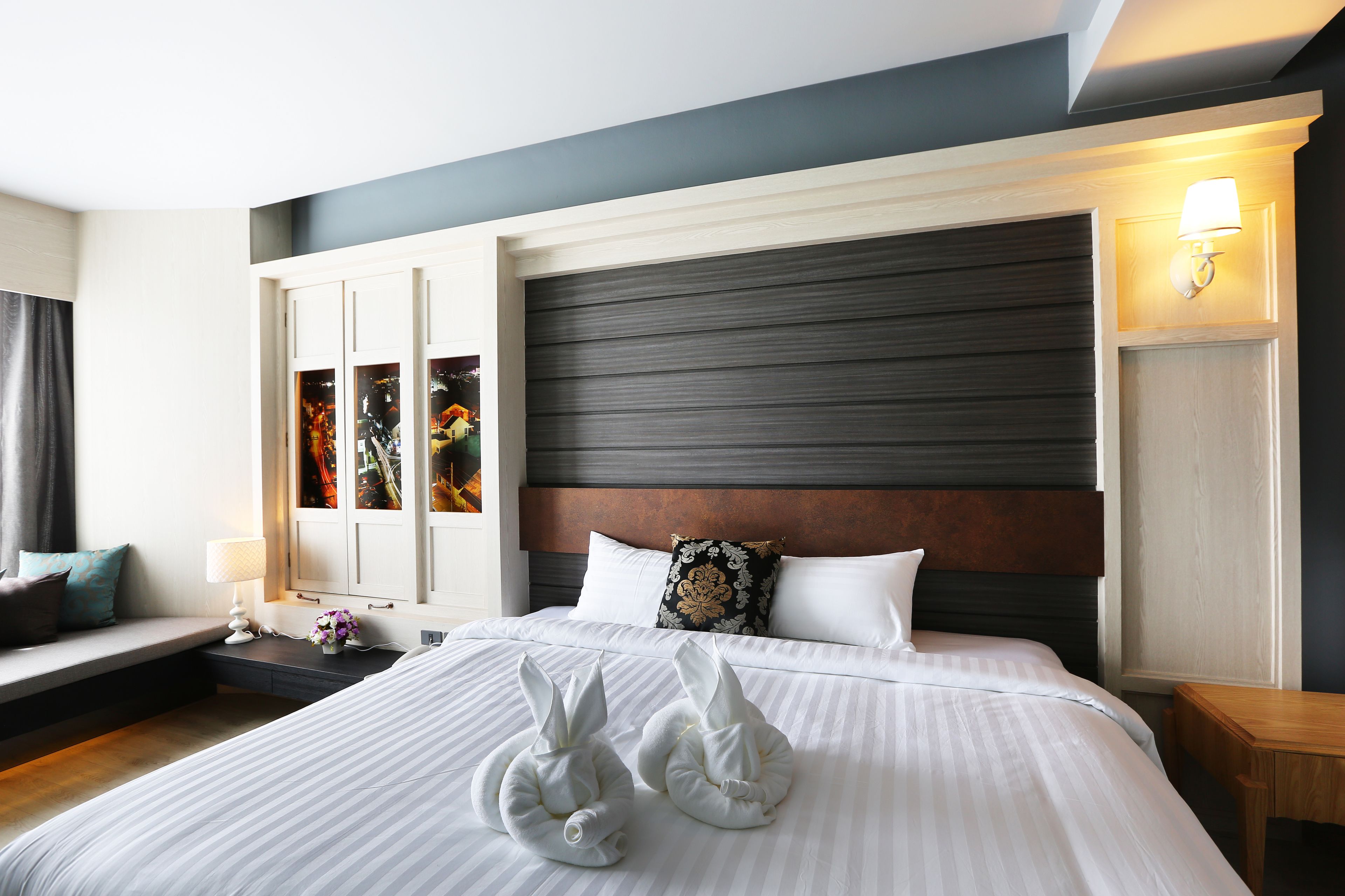 A modern hotel room with a large bed, a wooden headboard, a bedside table with a lamp, and a window with curtains.