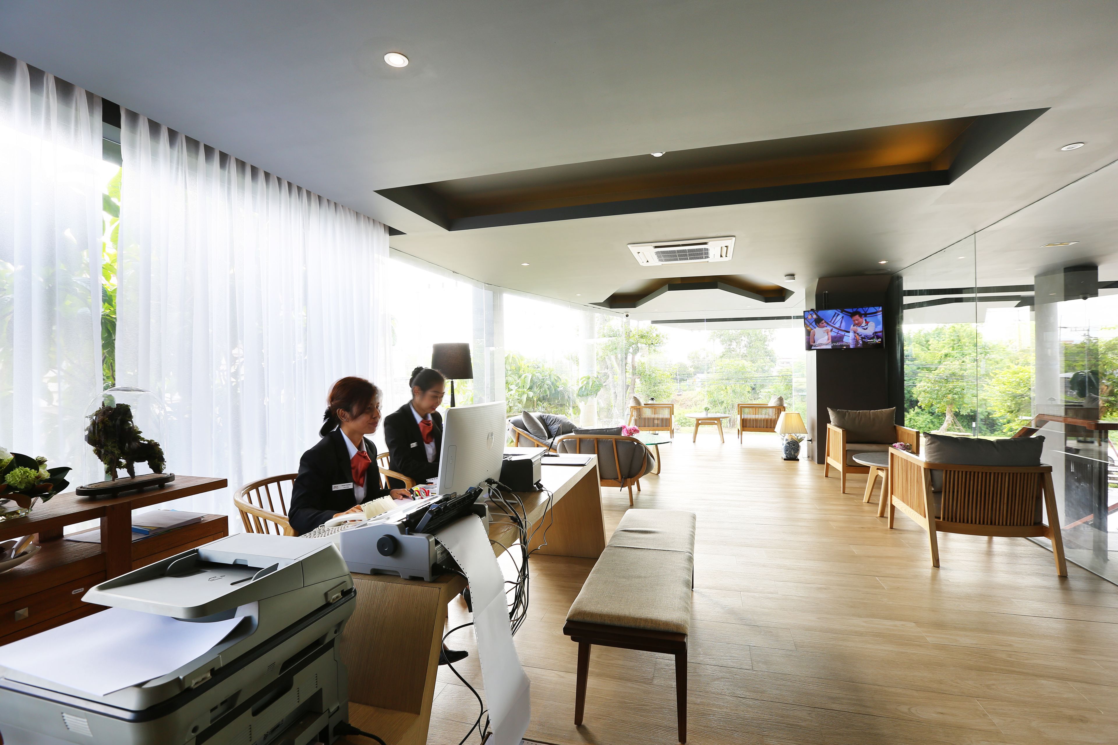 A modern office space with a large desk, computer, and printer. There are several chairs and a bench. A television is mounted on the wall.