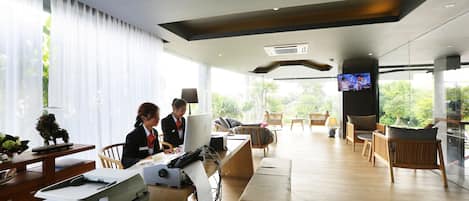 A modern office space with a large desk, computer, and printer. There are several chairs and a bench. A television is mounted on the wall.