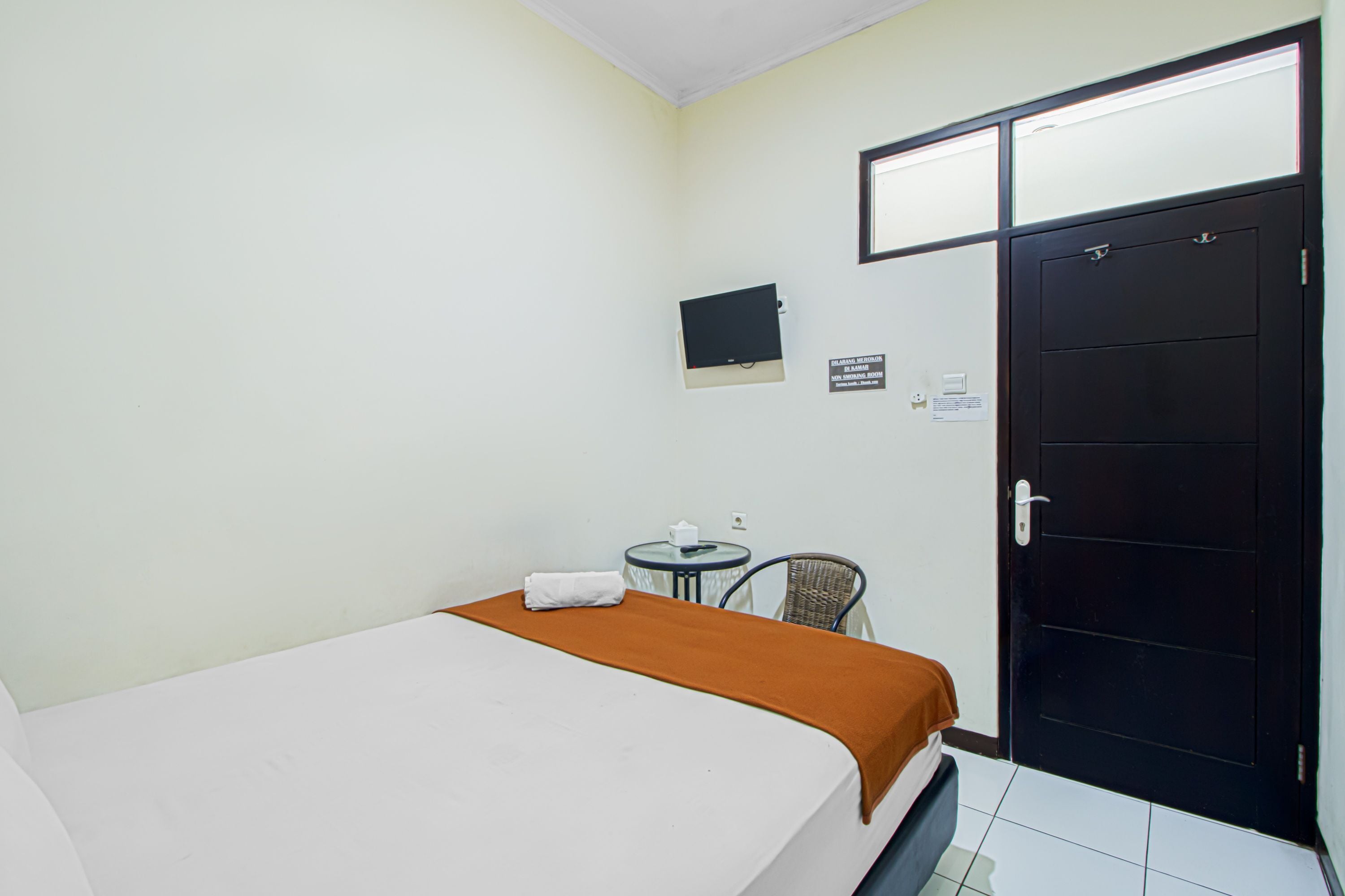 A hotel room with a bed, a small table with a chair, a TV mounted on the wall, and a black door.