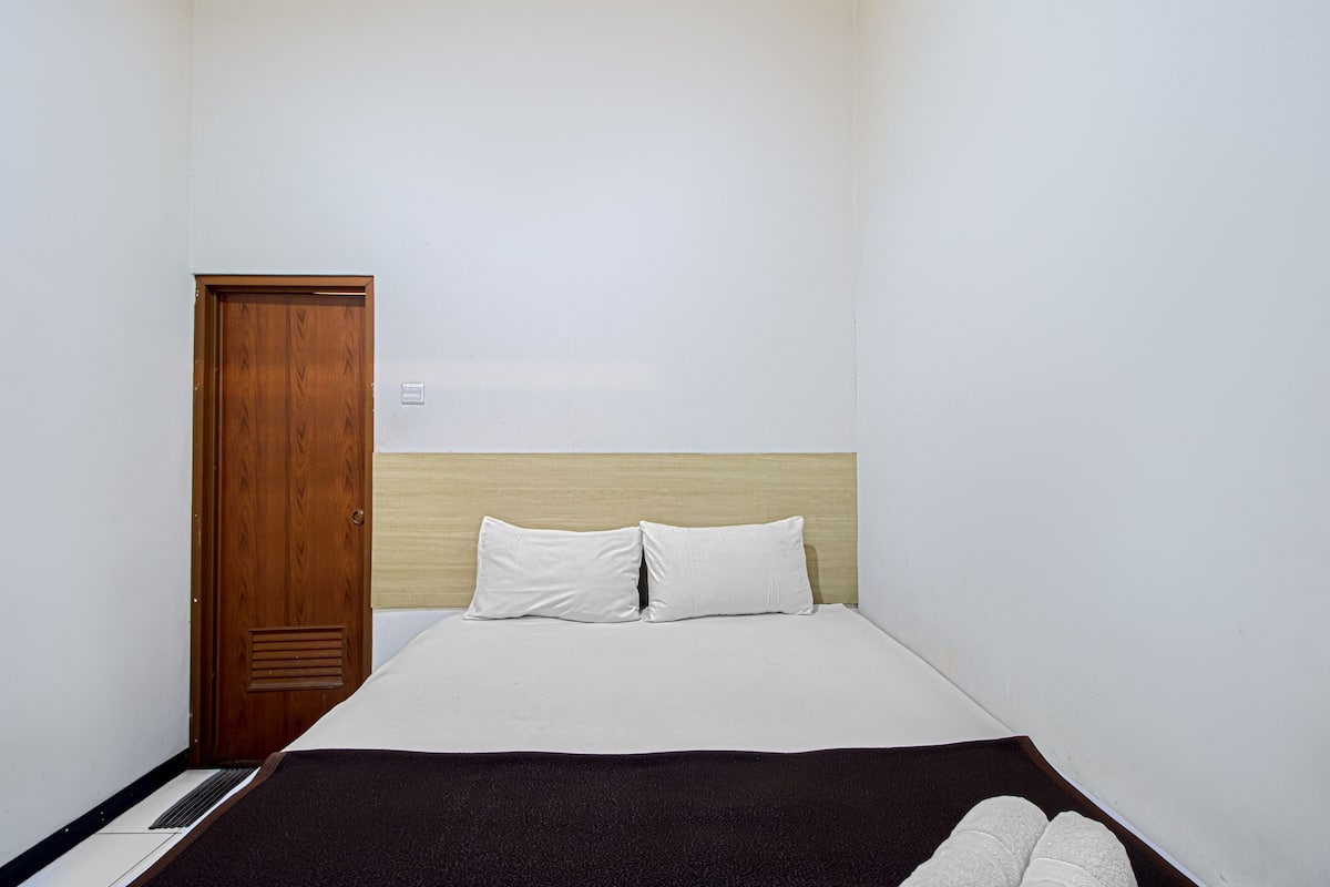 Ein kleiner, sauberer Raum mit Holztür, einem Bett mit weißer Bettwäsche und einer dunkel gefärbten Tagesdecke.