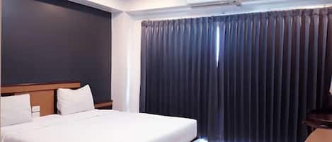 A modern hotel room with a large bed, a wooden headboard, a built-in air conditioning unit, and a window with dark curtains.
