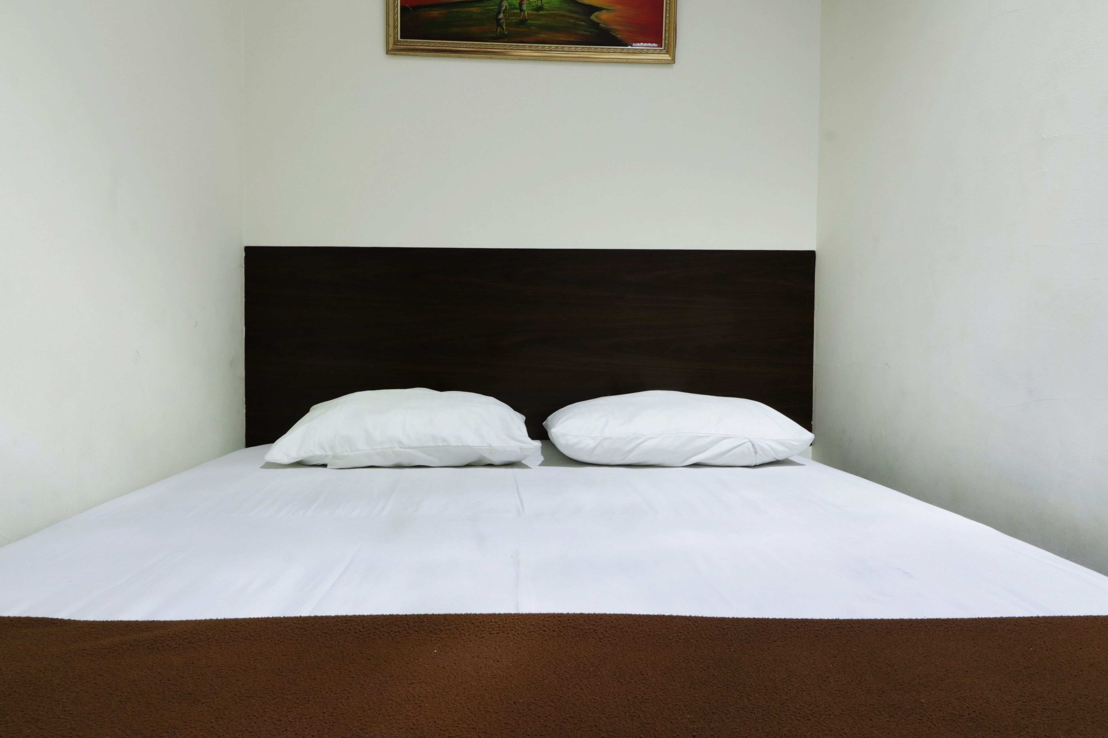 Una cama con ropa de cama blanca y un cabecero oscuro, un cuadro encima y una alfombra marrón al pie de la cama.