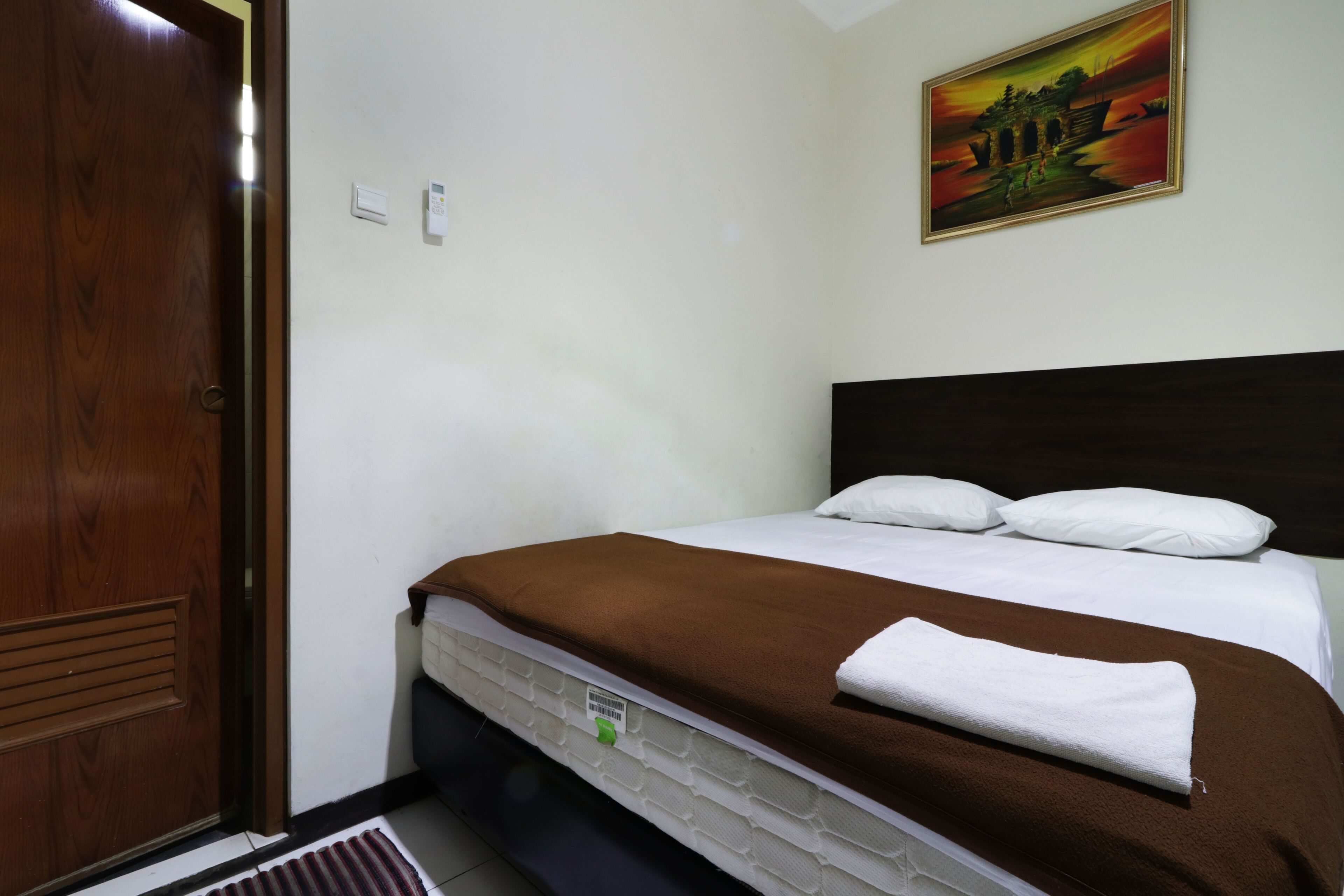 A hotel room with a bed, a wooden door, a painting on the wall, and a thermostat.