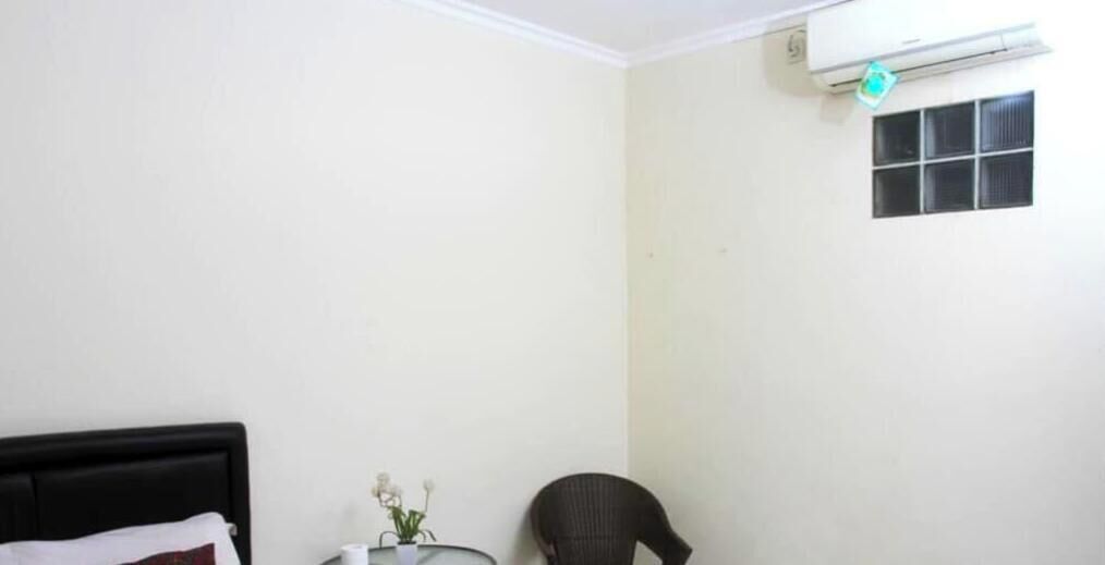 A room with a bed, a chair, a small table with a vase, and a window with blinds.