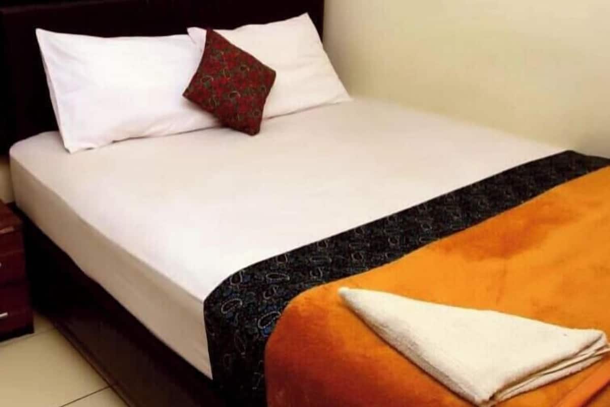 Ein ordentlich bezogenes Bett mit weißer und gemusterter Bettwäsche, einem braunen Kopfkissen und einem gefalteten weißen Handtuch auf einer orangefarbenen Decke.