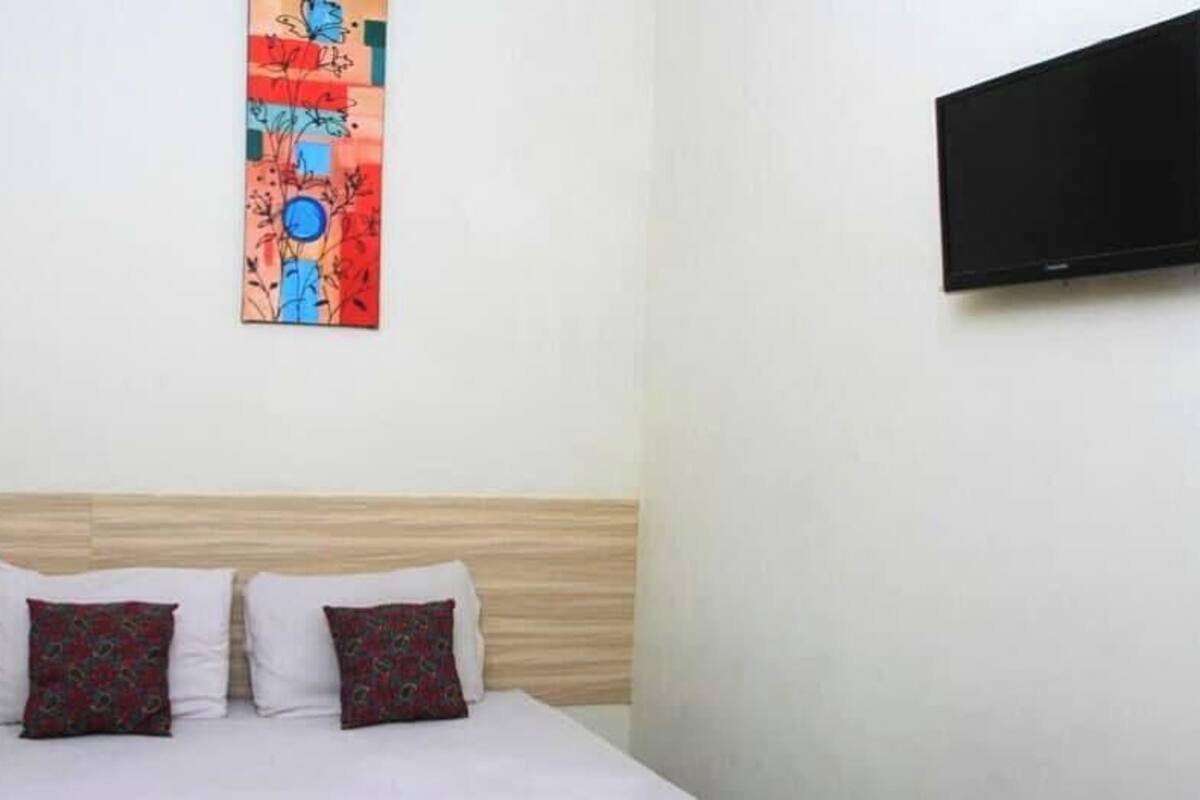 Ein Hotelzimmer mit einem Bett, zwei Kissen, einem an der Wand montierten Fernseher und einem farbenfrohen abstrakten Gemälde.