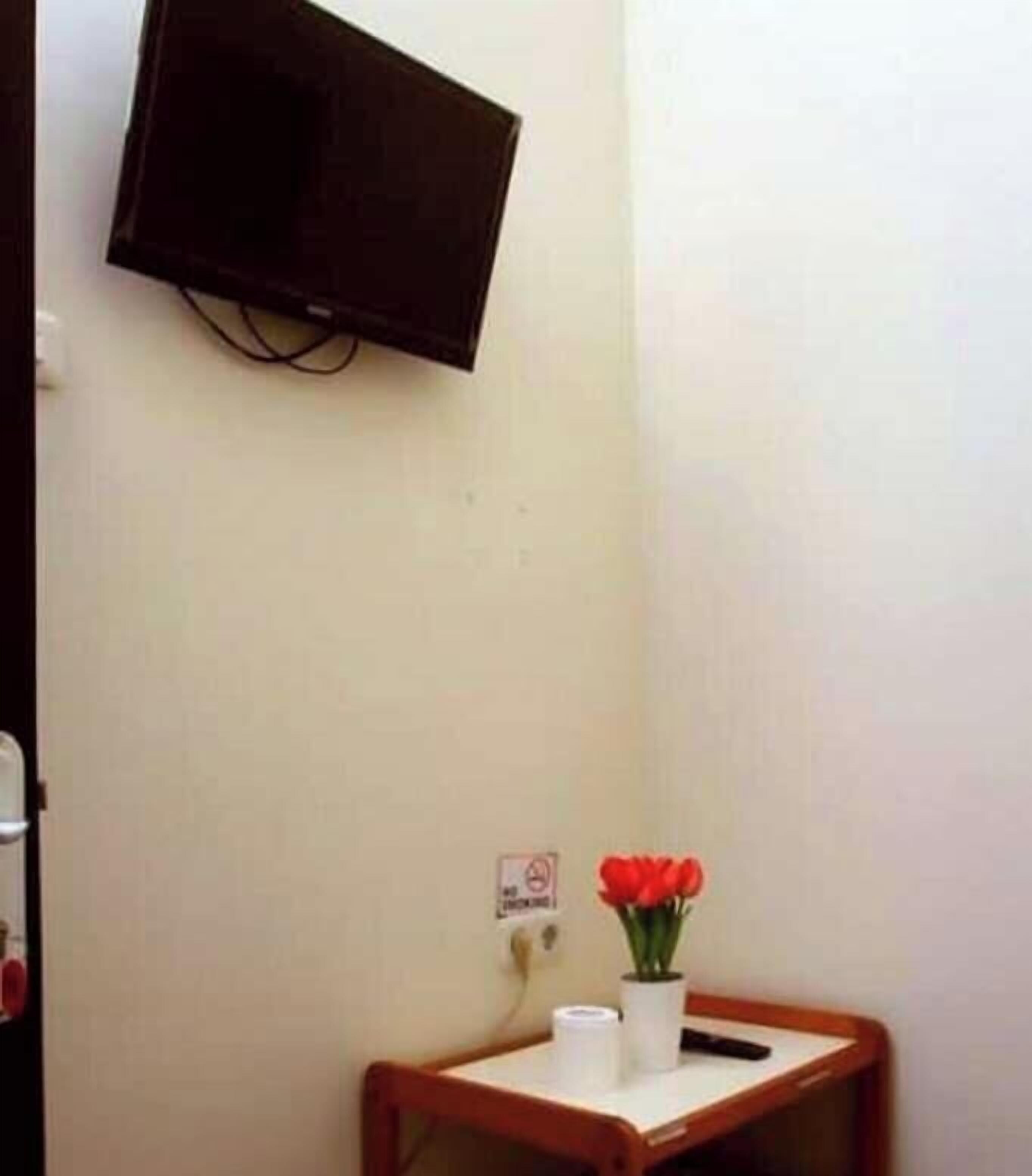 A small room with a television mounted on the wall, a wooden side table with a vase of red flowers, a roll of toilet paper, and a power outlet with a plug.