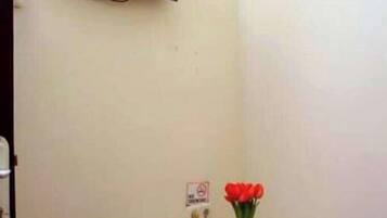 A small room with a television mounted on the wall, a wooden side table with a vase of red flowers, a roll of toilet paper, and a power outlet with a plug.