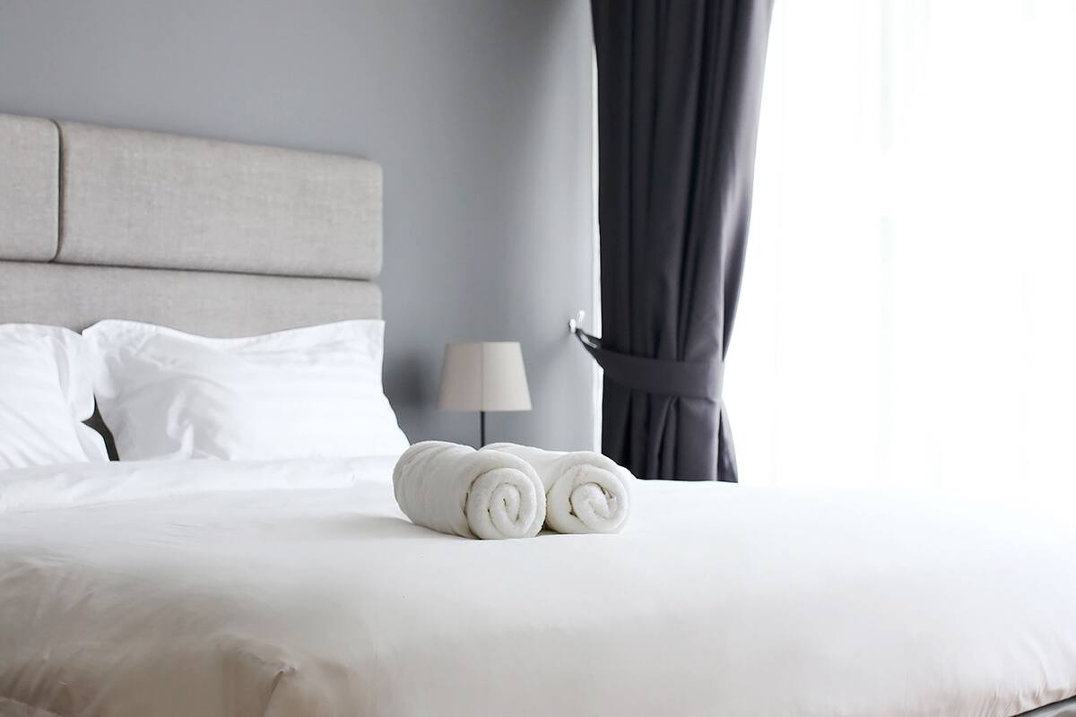 A neatly made bed with white linens and rolled towels, a bedside lamp, and a window with curtains.