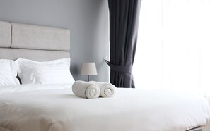 A neatly made bed with white linens and rolled towels, a bedside lamp, and a window with curtains.