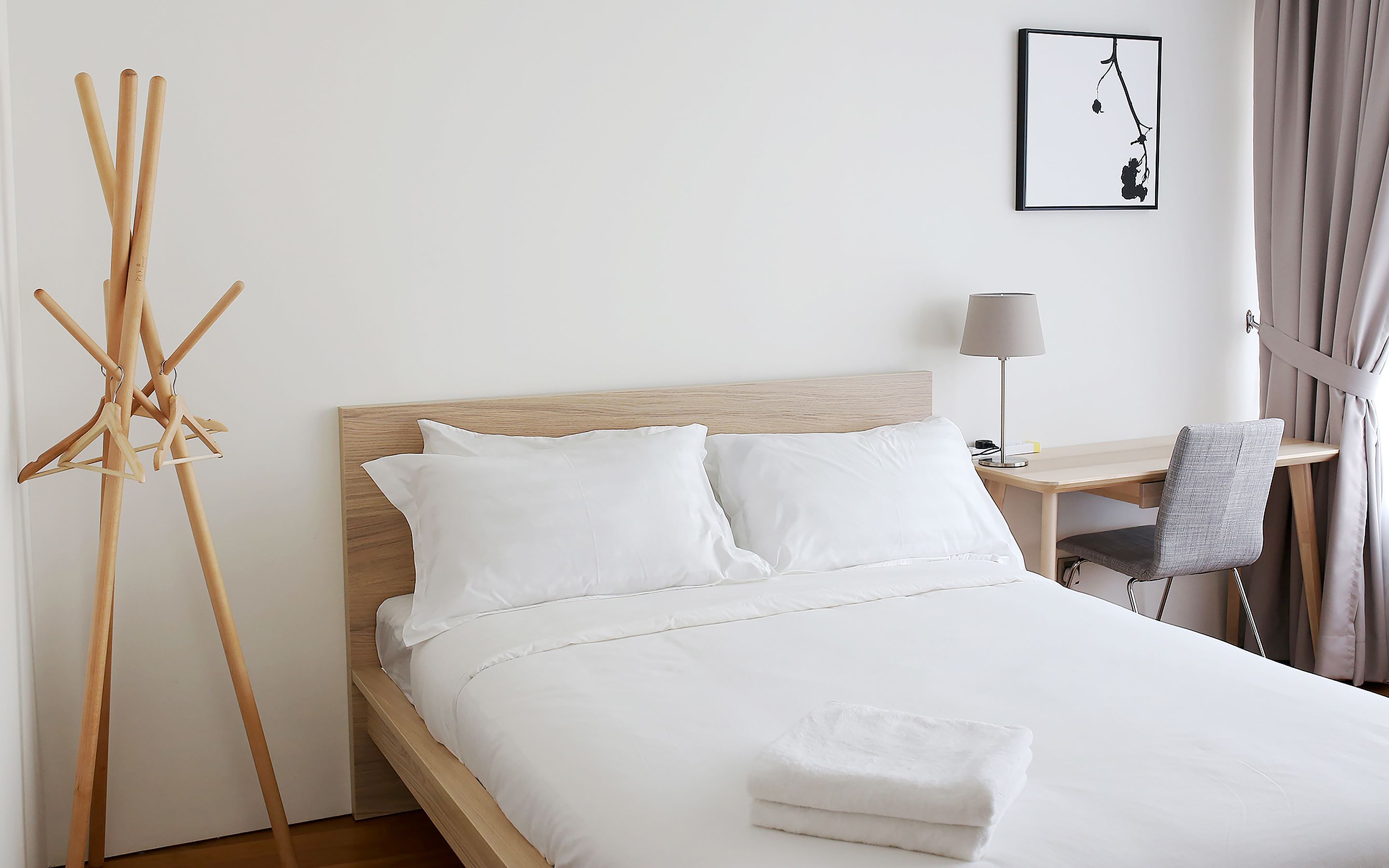 Una cama bien hecha con sábanas blancas, un cabecero de madera, una mesita de noche con una lámpara y un perchero con perchas.