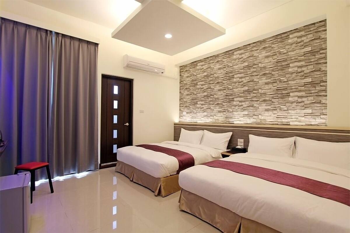 A hotel room with two beds, a desk, a chair, and a stone wall.