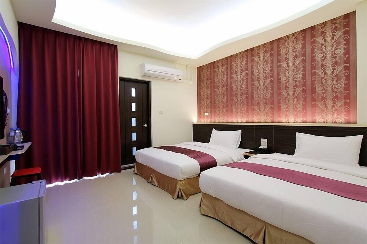 A hotel room with two beds, a desk, a chair, and a large window with curtains.