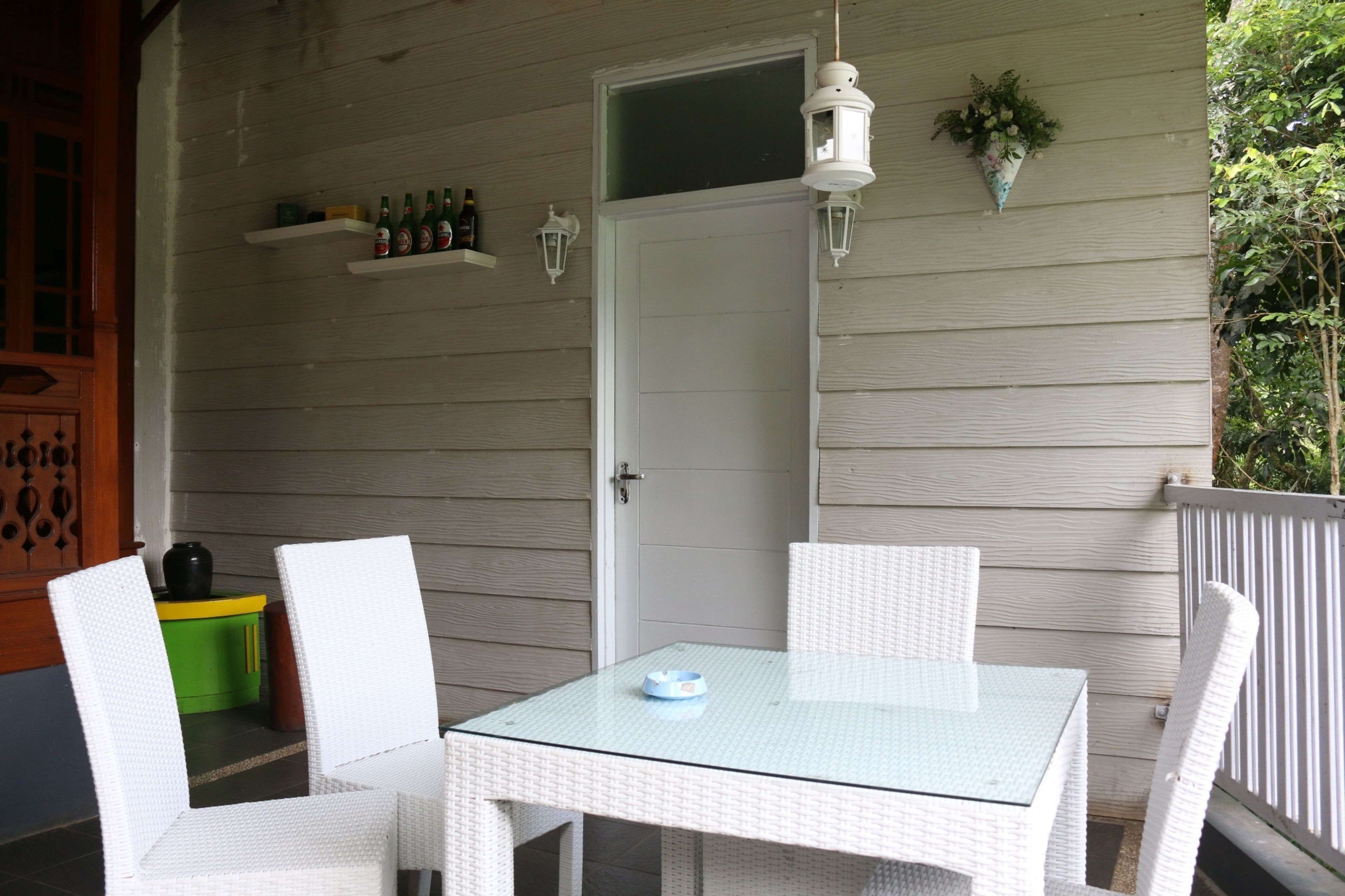 Varanda coberta com mesa de vidro e cadeiras de vime branco. Prateleiras com garrafas e um candeeiro na parede.