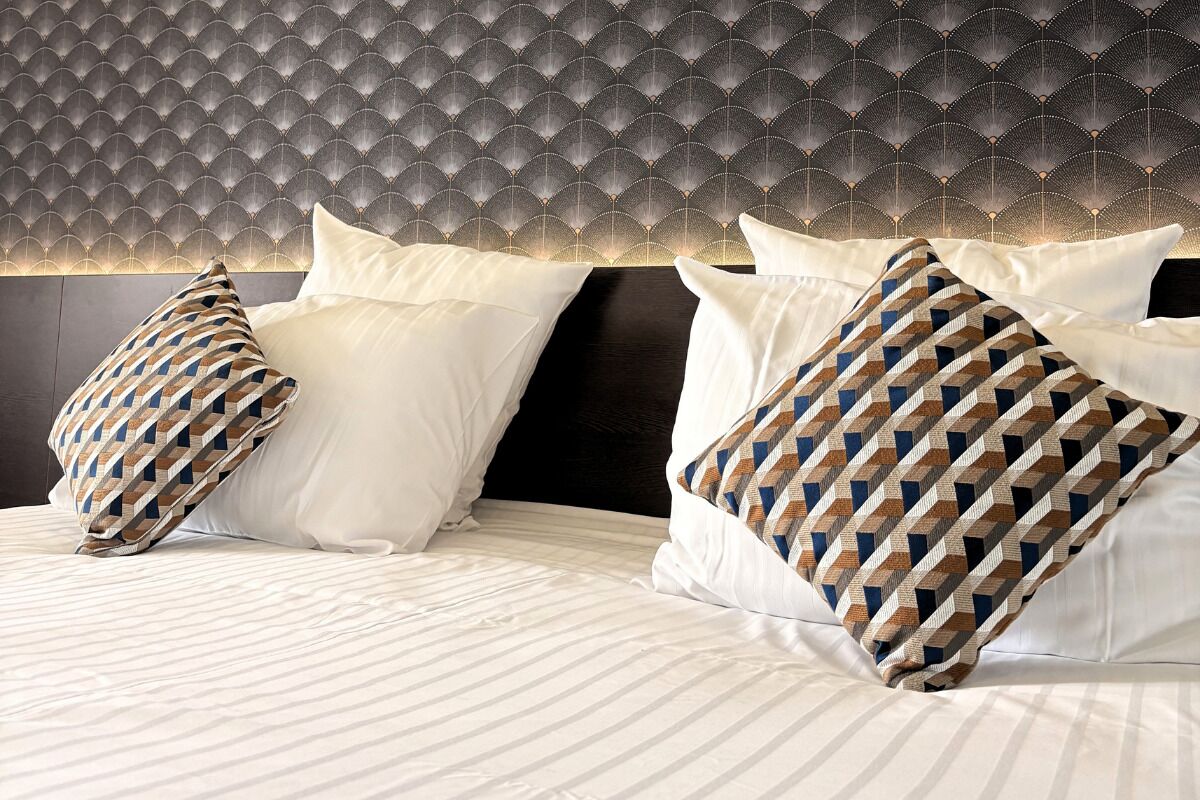 A hotel room with a patterned headboard, white bedding, and decorative pillows.