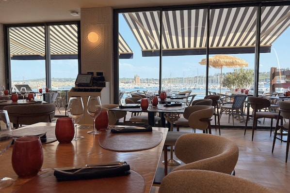 A restaurant with large windows, tables, chairs, and a view of the marina.
