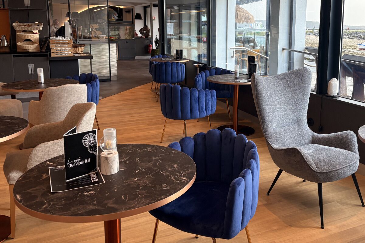A modern interior with round tables, blue and grey upholstered chairs, and a visible kitchen area.