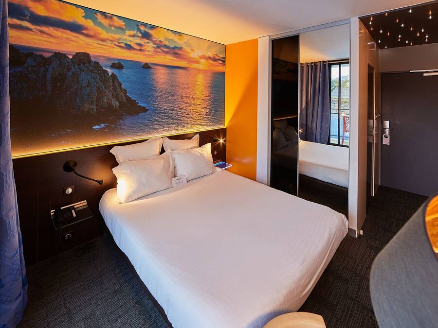 A hotel room with a large bed, a nightstand, a desk, and a view of the sea through a window.