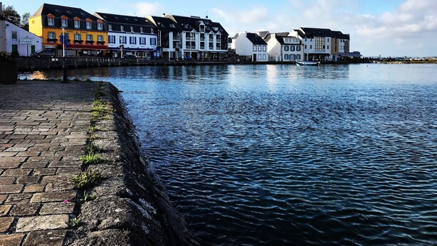 A waterfront hotel with a brick walkway, a row of colorful buildings, and a calm river.