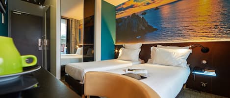 A hotel room with two beds, a desk with a green mug, a painting of a sunset, and a nightstand with a lamp.