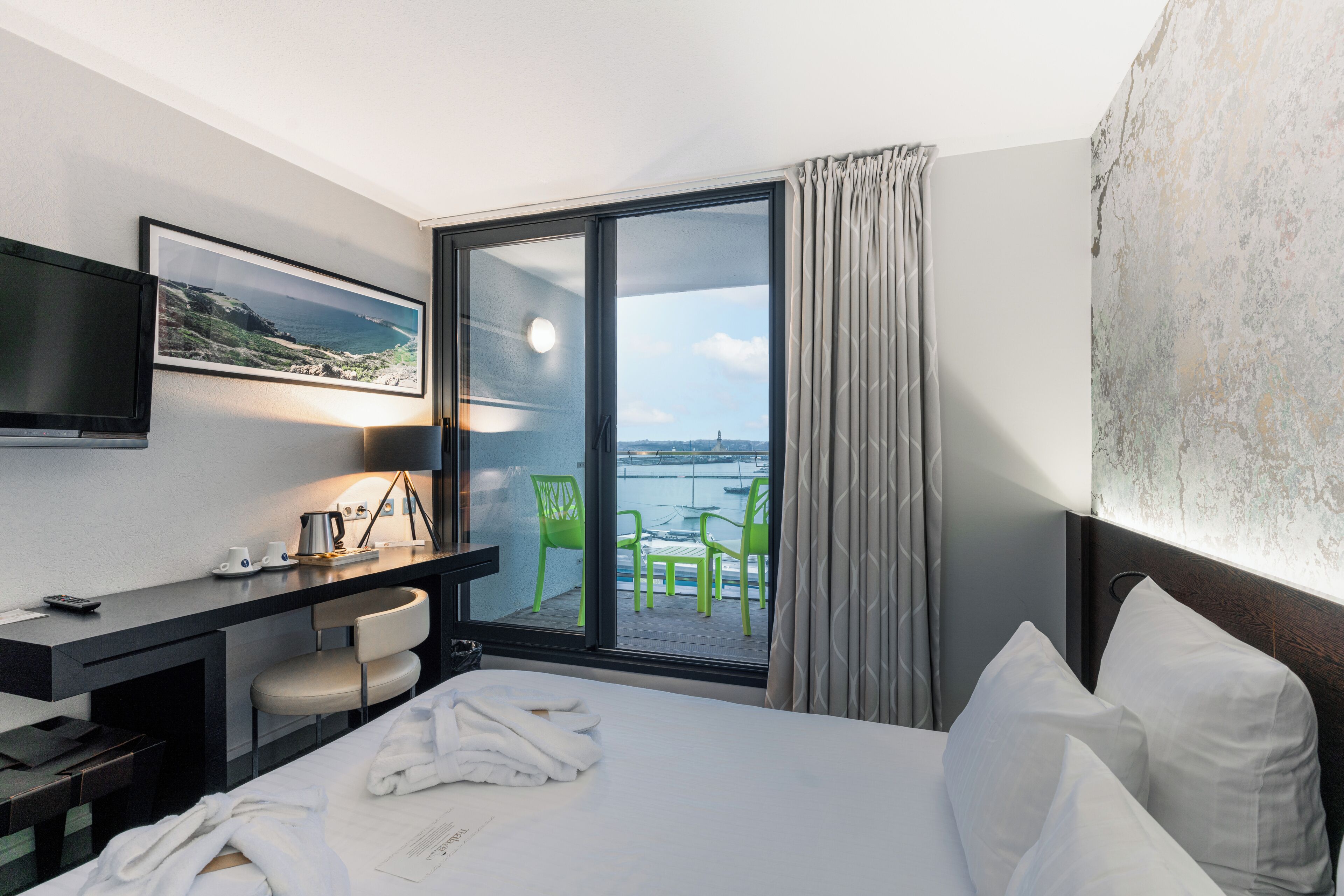 A hotel room with a bed, a desk with a chair, a TV, and a balcony with chairs.