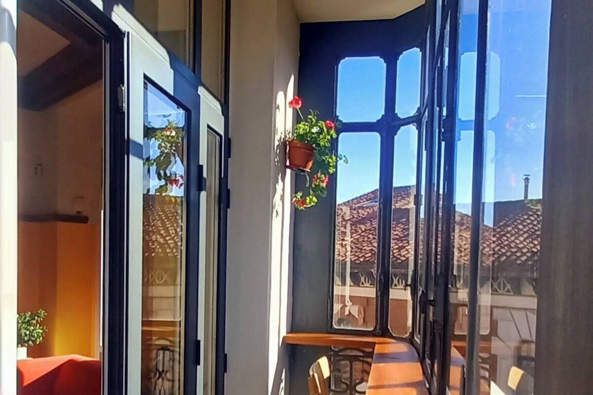 A narrow balcony with a wooden table and chairs, large windows, and hanging flower baskets.