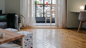 Et værelse med trægulv, et bord med en hvid bænk, en stol, en sofa med et grønt pled og en balkon med en plante og en rød kop.
