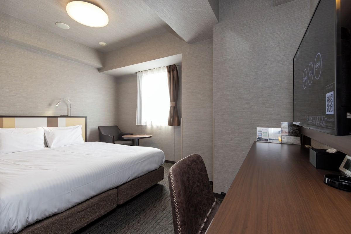 Habitación de hotel con cama, escritorio, silla, televisor y ventana con cortinas.