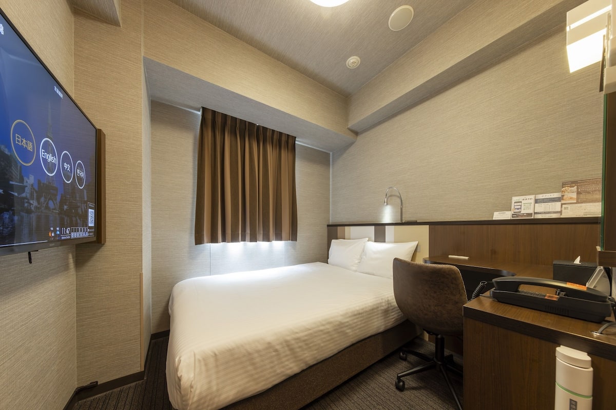 Habitación de hotel con cama, escritorio con teléfono, silla, televisor montado en la pared y ventana con cortinas.