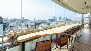 A modern bar with a long counter and multiple stools, overlooking a cityscape through large windows.