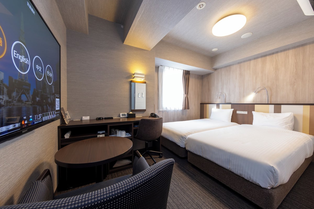 Habitación de hotel con dos camas, un escritorio, una silla, un televisor y una ventana con cortinas.