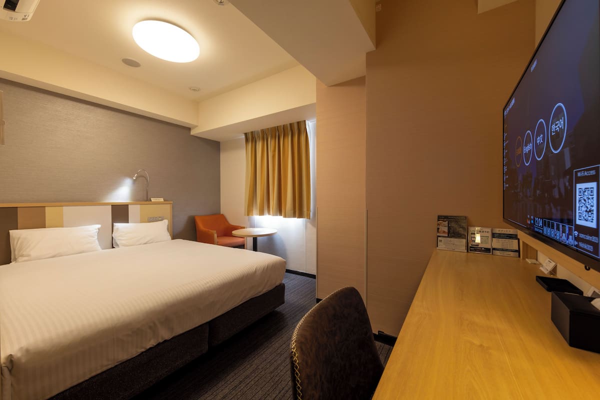 Habitación de hotel con una cama grande, un escritorio, una silla, una mesita y una pantalla de televisión mostrando información.