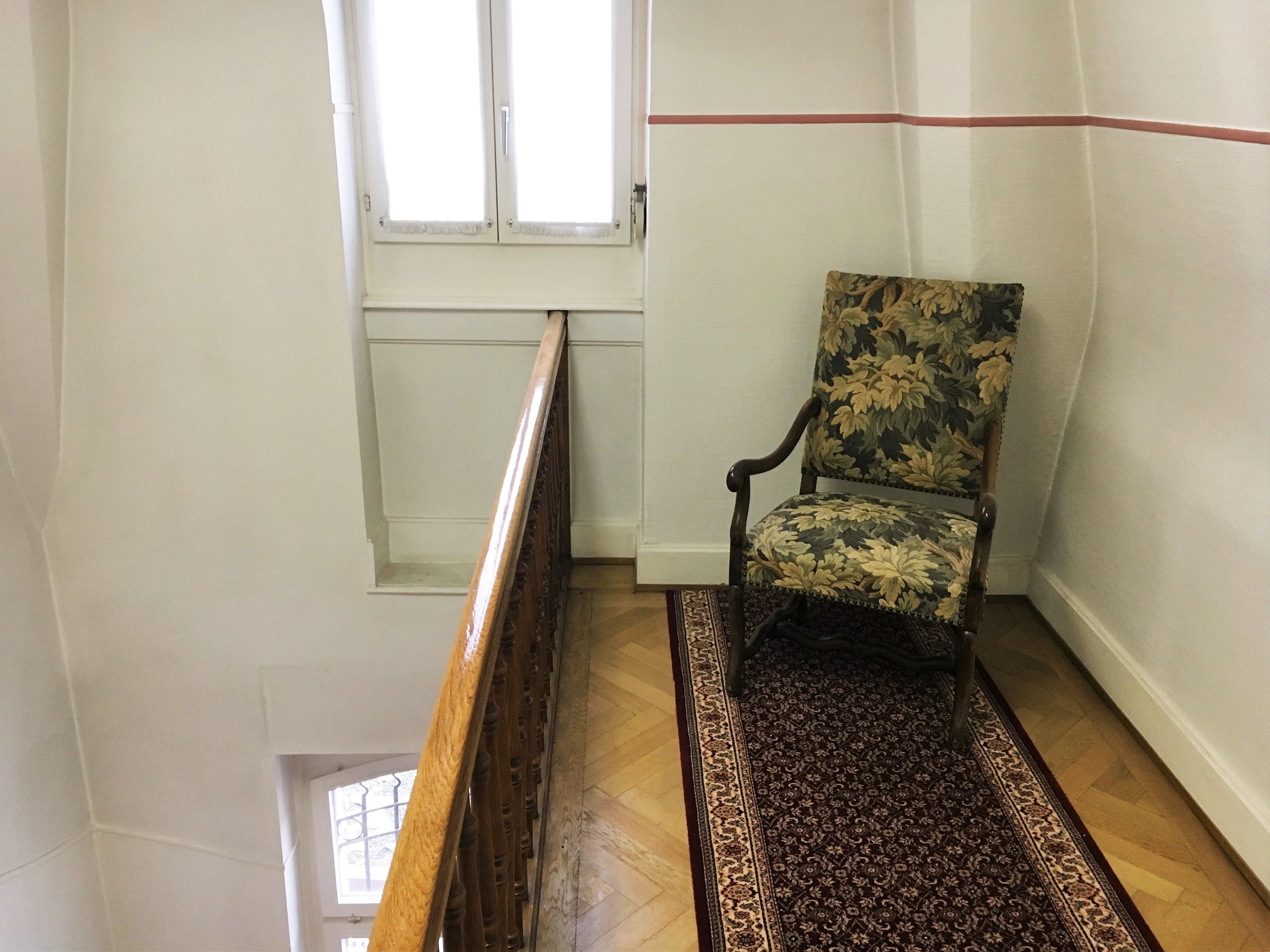 A chair in a corner of a room with a patterned rug, wooden floor, and a window with blinds.
