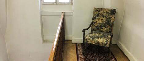 A chair in a corner of a room with a patterned rug, wooden floor, and a window with blinds.