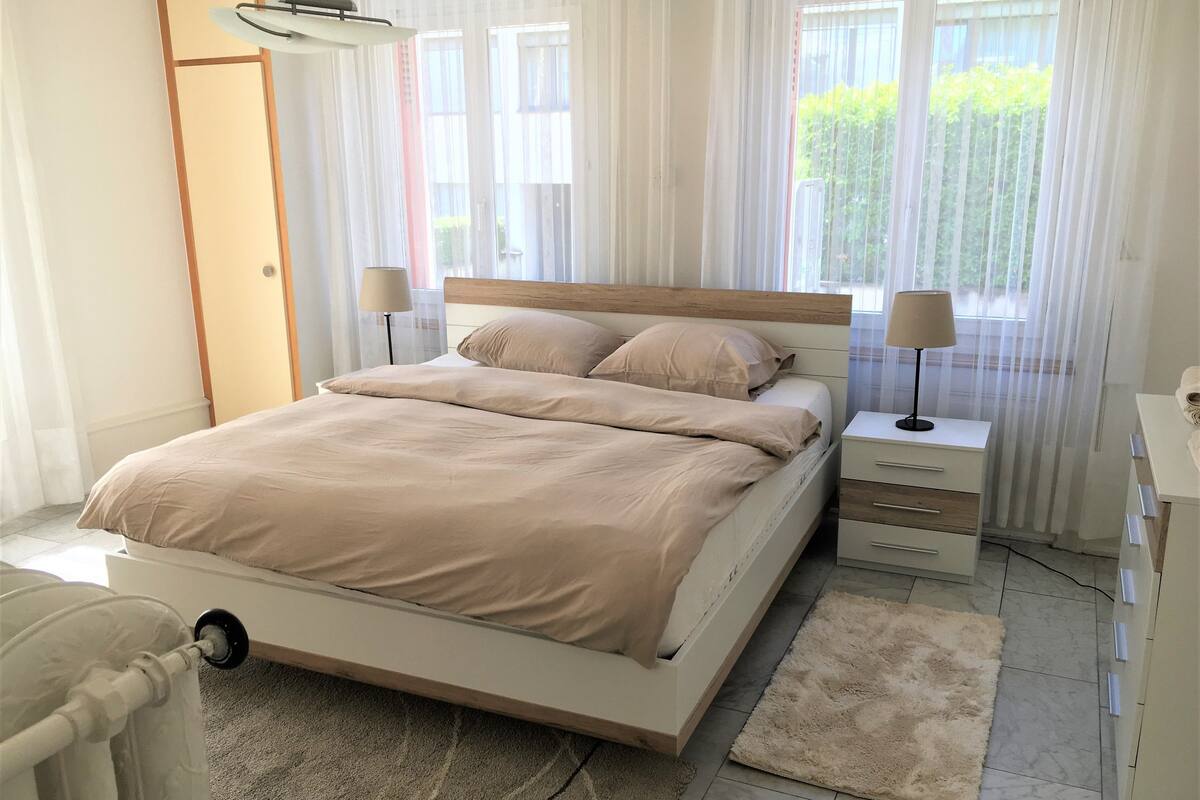 Une chambre à coucher comprenant un lit, des tables de chevet, une commode, une chaise et une vue sur la verdure à l’extérieur.