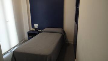 A small, narrow hotel room with a single bed, a nightstand, and a wall-mounted telephone.