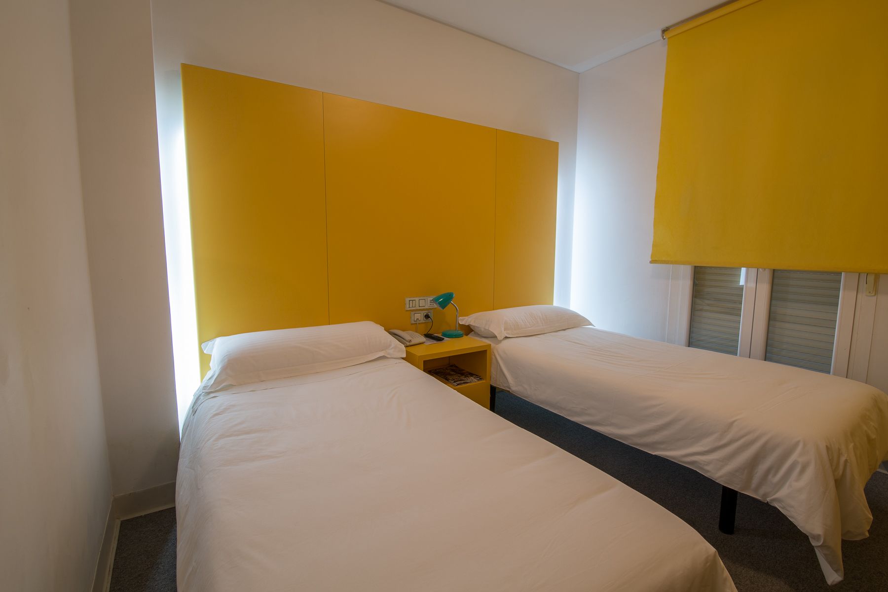 A hotel room with two single beds, a small bedside table with a lamp, and a window with a yellow blind.