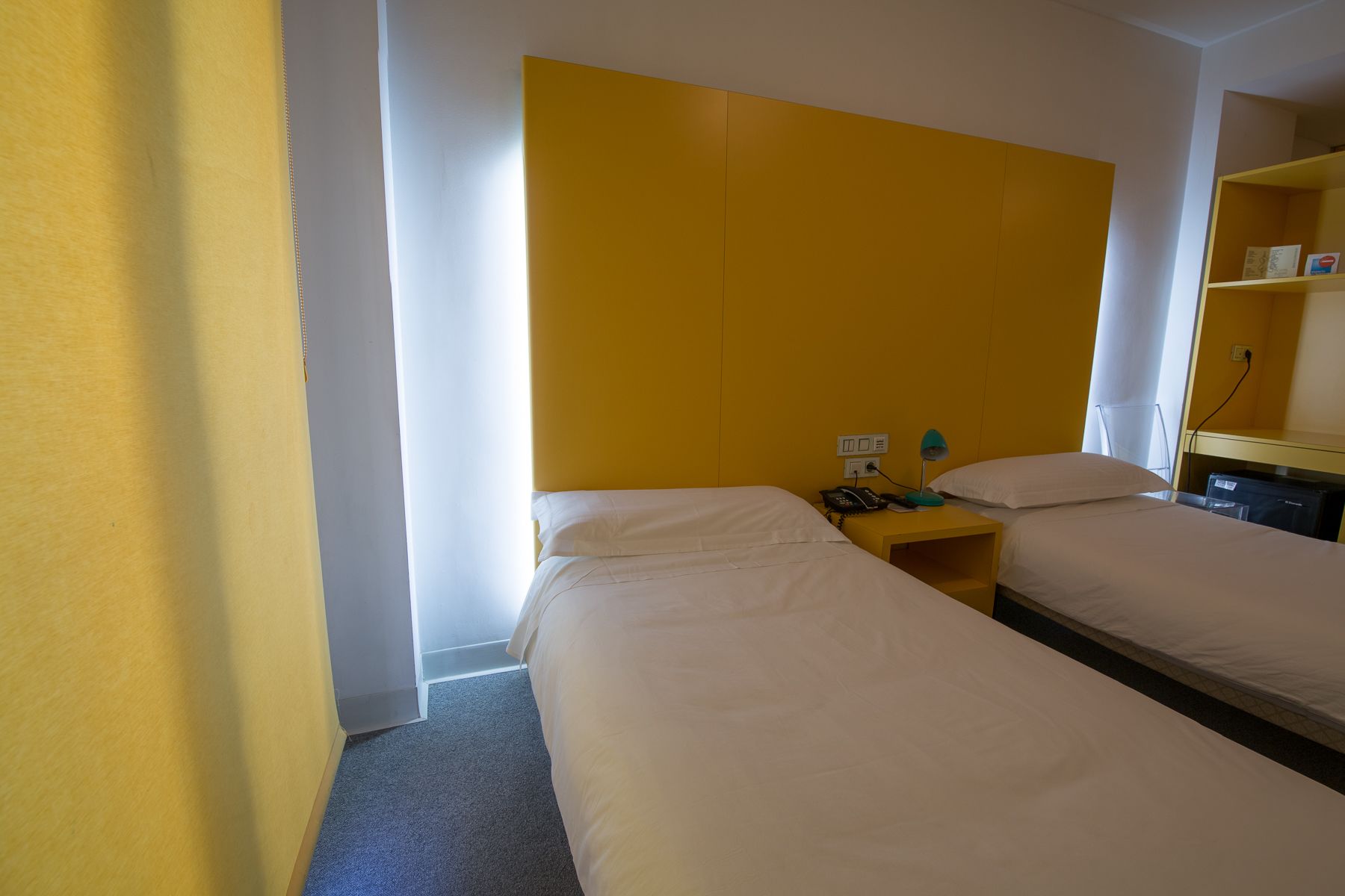 A hotel room with two single beds, a bedside table with a lamp, a phone, and a shelf with a book.