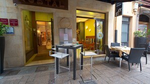 Een hotelentree met een bordje waarop 'HOSTAL' staat, buiten zitplaatsen met tafels en stoelen, en een bloempot links.