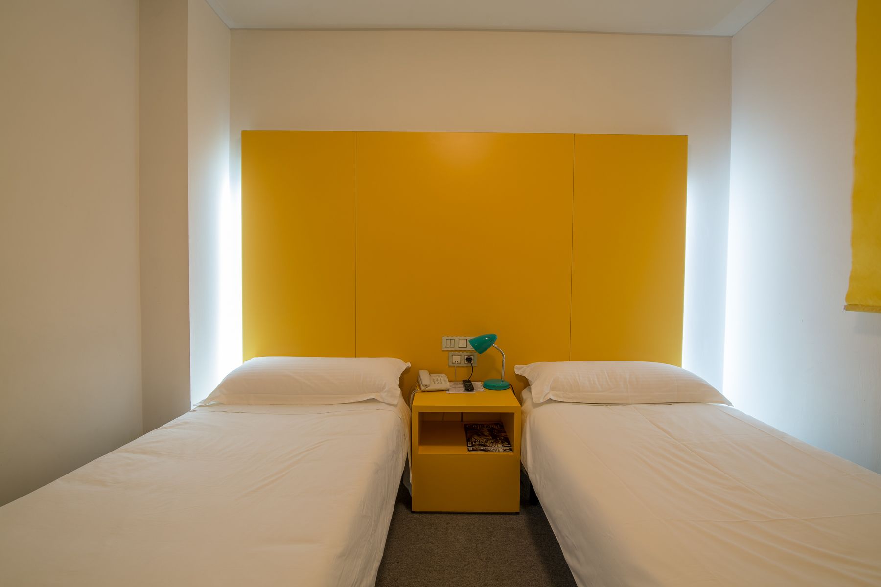 Una habitación con dos camas, un cabecero amarillo, una mesita de noche con lámpara y teléfono.
