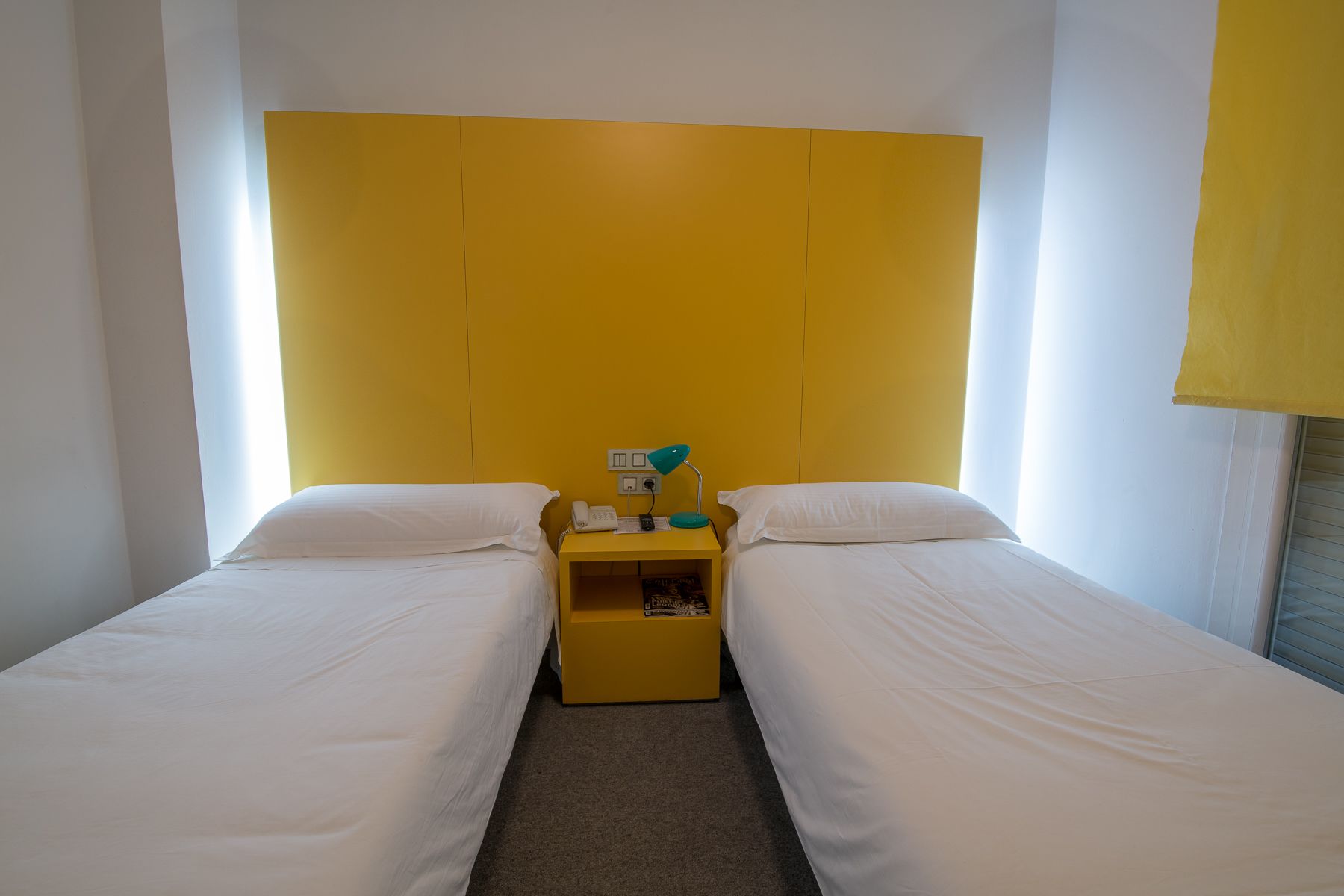 Une chambre d’hôtel avec deux lits simples, une petite table de chevet jaune avec une lampe et une fenêtre avec des rideaux.