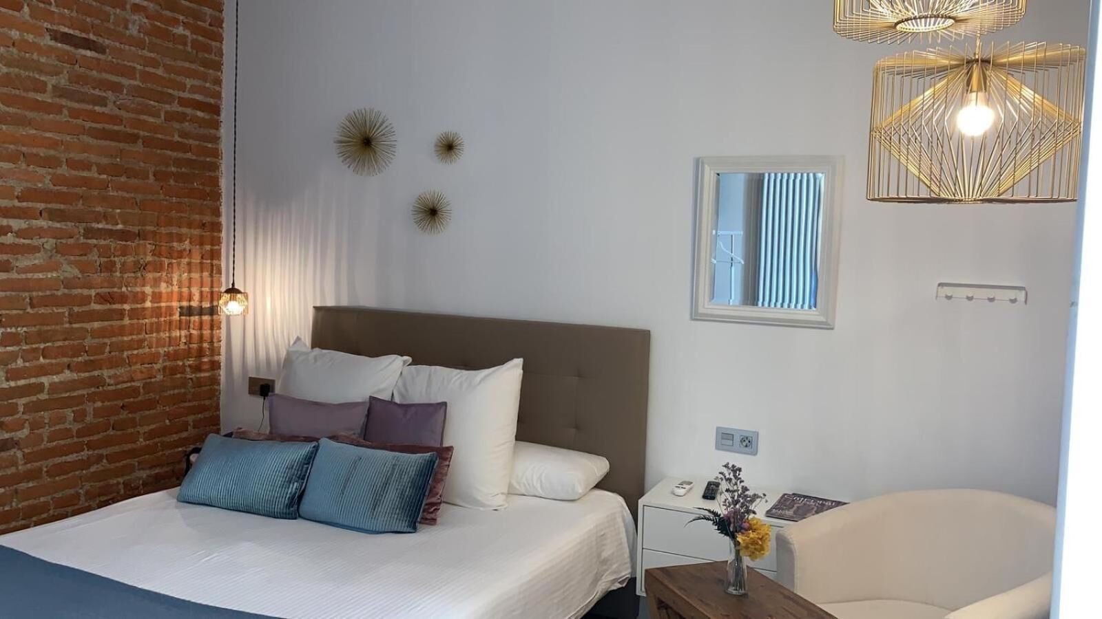 A bedroom with a brick wall, a bed with multiple pillows, a nightstand with a vase, and a hanging light fixture.