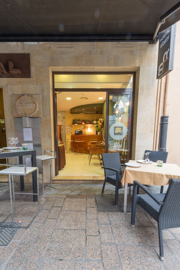 An outdoor seating area with tables and chairs, a bar visible through the entrance, and a sign indicating a hostel. - Hostal Albany Ancha (León)