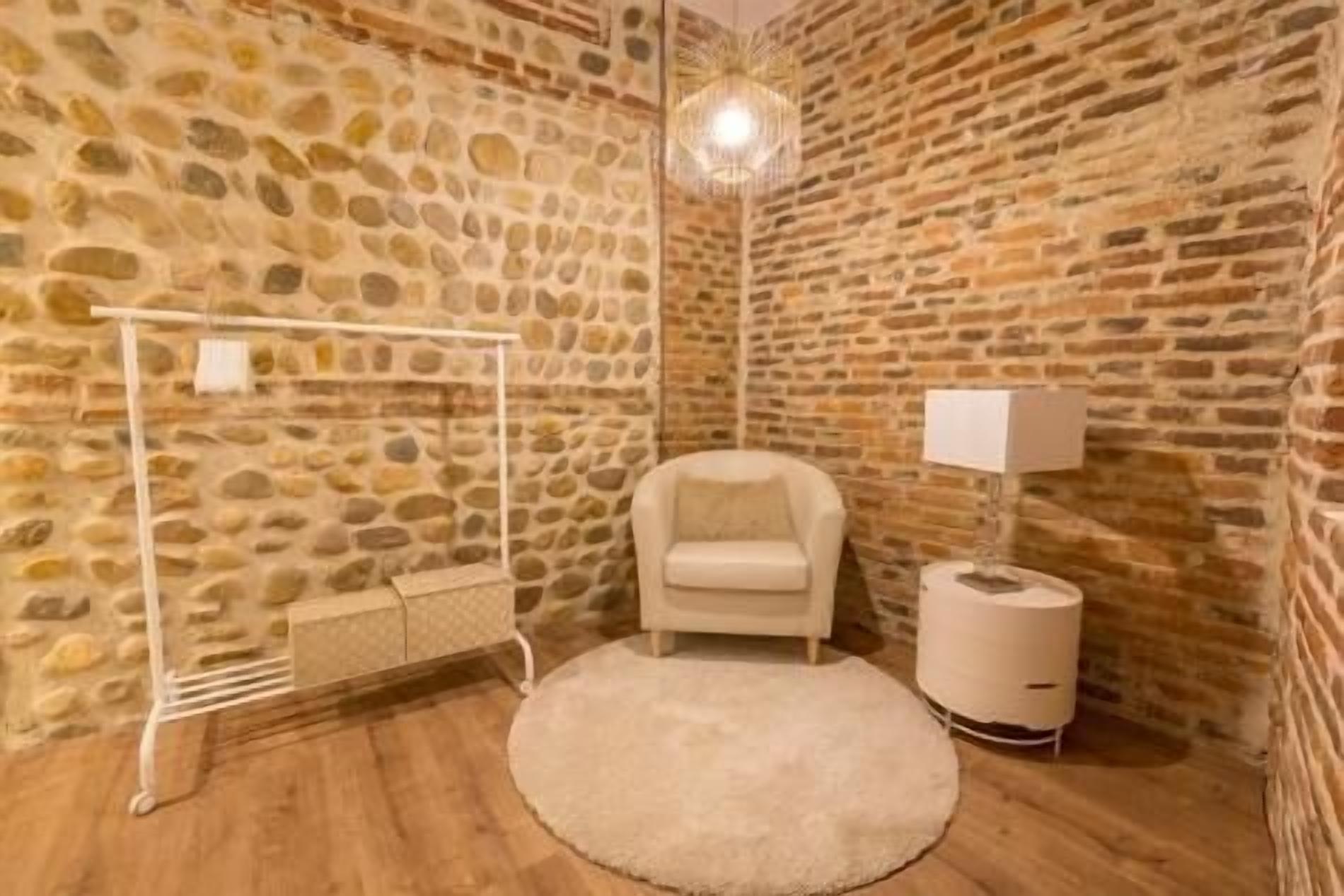 A room with stone walls, a round rug, a white chair, a lamp, and a hanging light fixture.