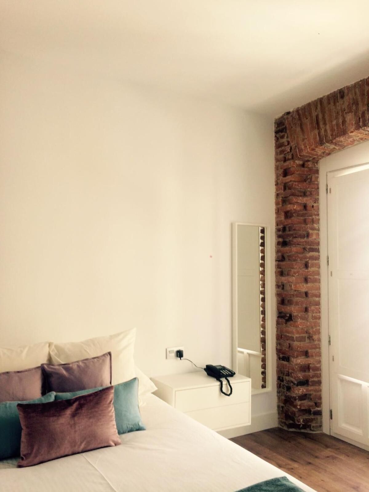 Un dormitorio con cama, almohadas, mesita de noche con teléfono, espejo y pared de ladrillo.