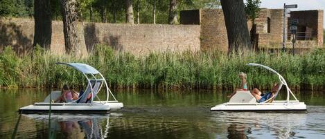 Two small boats with canopies, each carrying a person. The boats are on calm water, surrounded by tall grass and trees.