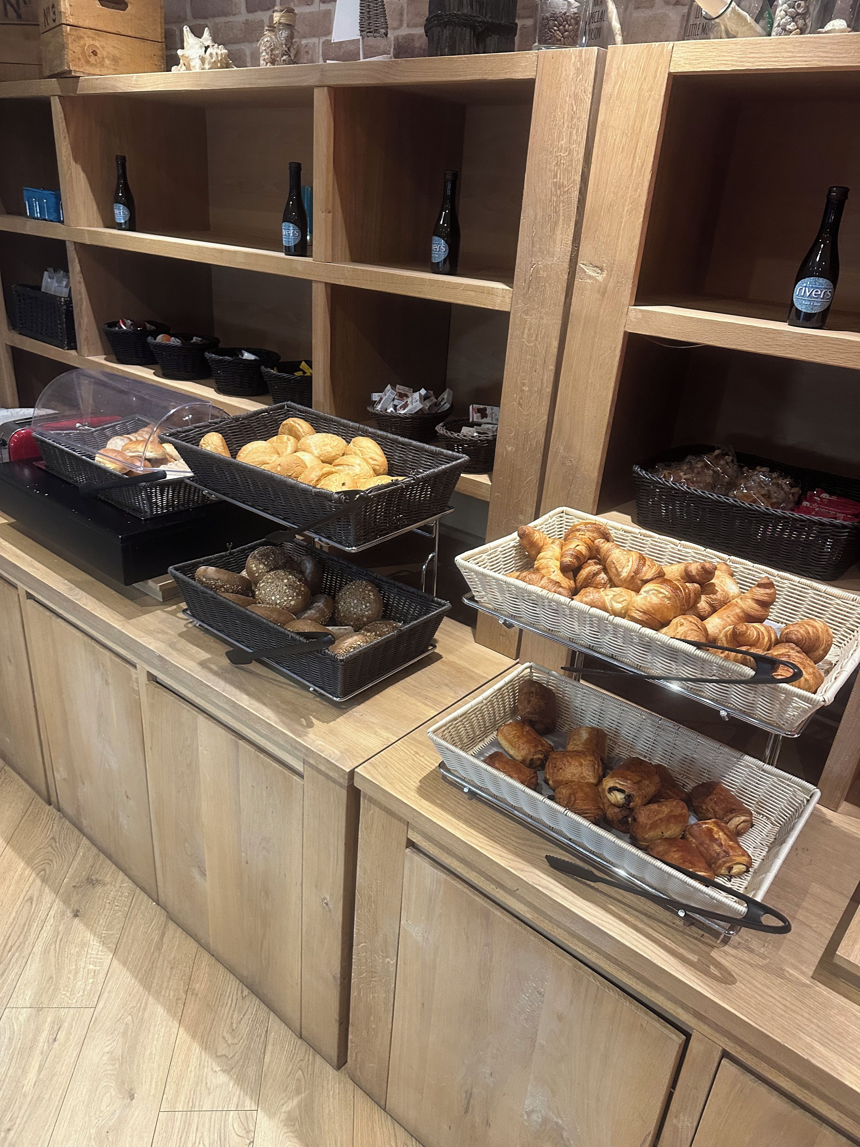 Een buffetgedeelte met verschillende manden gebak en brood, houten planken met flessen en een balie met een glazen vitrine.