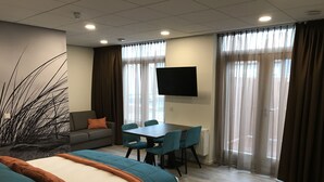 Een moderne hotelkamer met een bed, een sofa, een eettafel met stoelen, een flatscreen televisie en grote ramen met gordijnen.