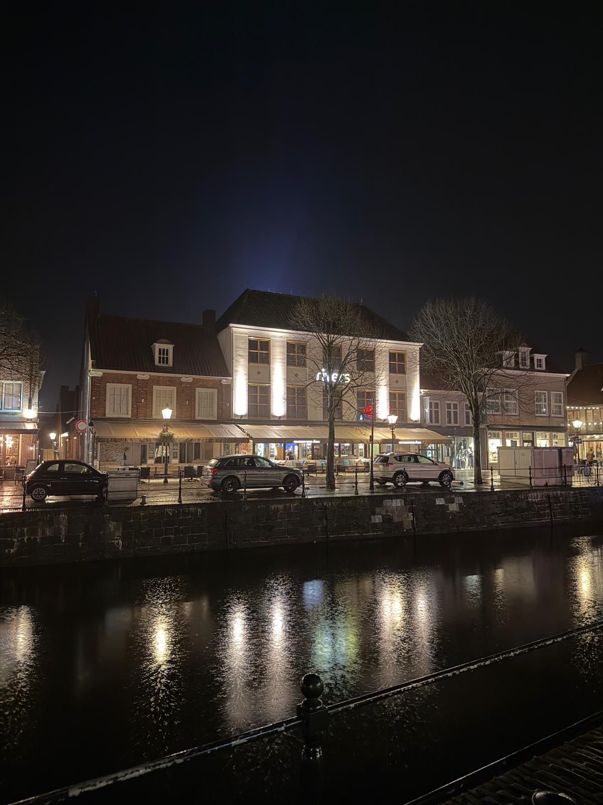 Een nachtzicht van een straat langs de rivier met gebouwen, een brug en geparkeerde auto's.